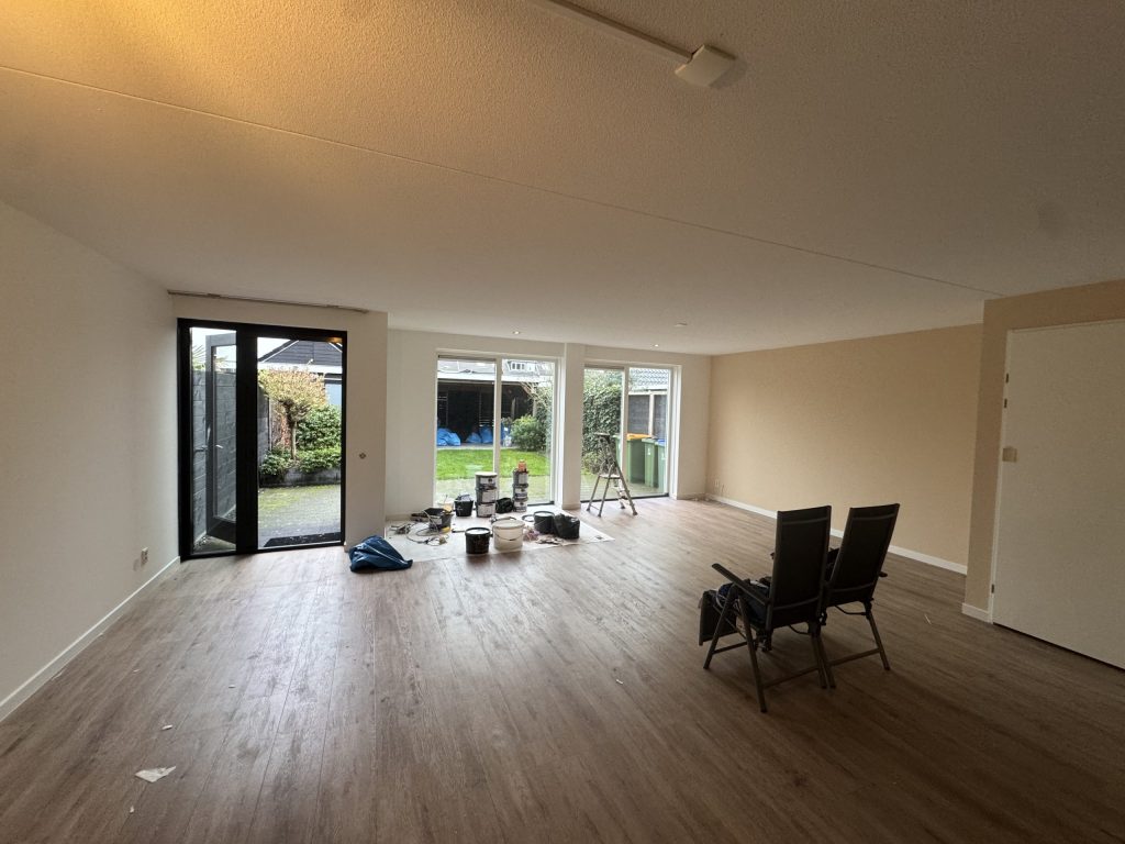 Ruime woonkamer met tuindeuren en een houten vloer, met verfemmers klaar voor de afwerking
