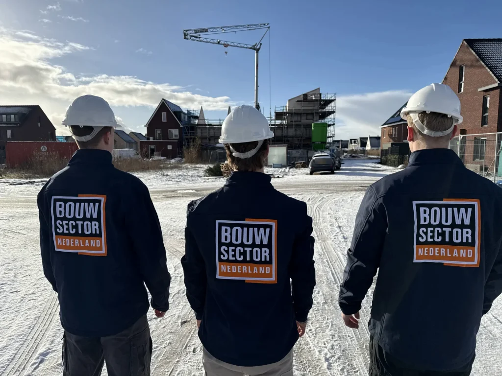 Drie vakmensen in donkerblauwe jassen met het logo van Bouwsector Nederland kijken naar een nieuwbouwwijk